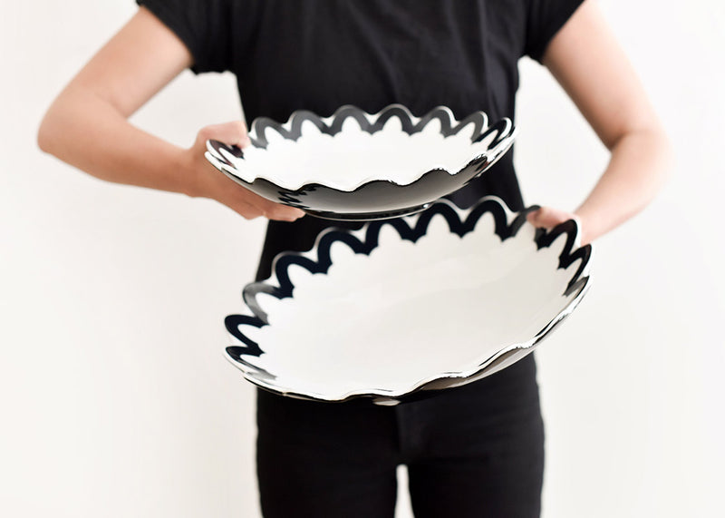 A person holding a Black Arabesque Scallop 11 Pasta Bowl, showcasing a black arabesque pattern on a white base. Perfect for serving and as stylish décor.