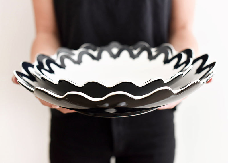 Person holding Black Arabesque Scallop 11 Pasta Bowl, showcasing bold arabesque pattern on white base. Versatile for serving and decor, pairs beautifully with 14-inch pasta bowl. Ideal for modern tablescapes.