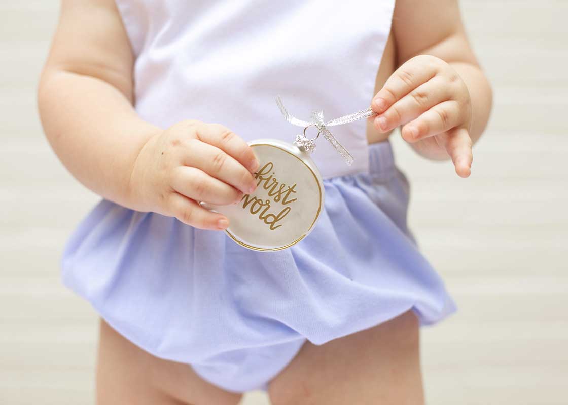 Front View of Child holding First Word Ornament from Baby's First Milestones in One Hand and Pulling on Ribbon.
