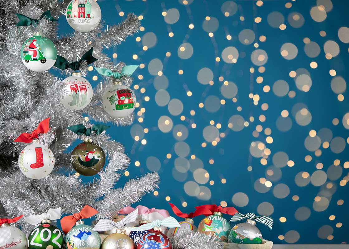 Front View of Christmas Ornaments Including Away We Go Design on Silver Tinsel Tree with Twinkle Lights in Background