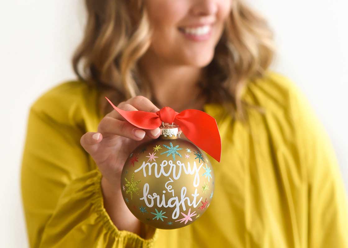 Front View of Woman Smiling and Holding Merry and Bright Stars Glass Ornament out in Front of Her