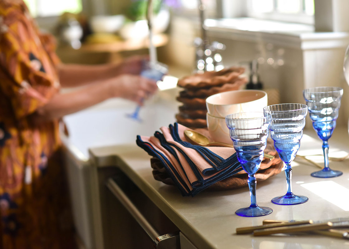 Front View of Woman Washing a Glass in a Kitchen Sink Next to Stacks of Ruffle Dinner Plates and Coordinating Fundamental Designs