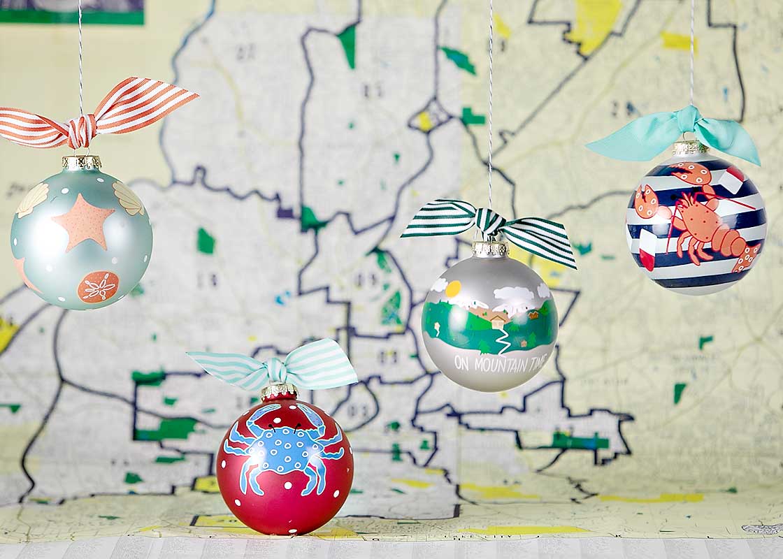 Front View of Glass Travel-themed Ornaments Including Crab, Lobster, Mountain, and Shell Designs Placed in Front of Map