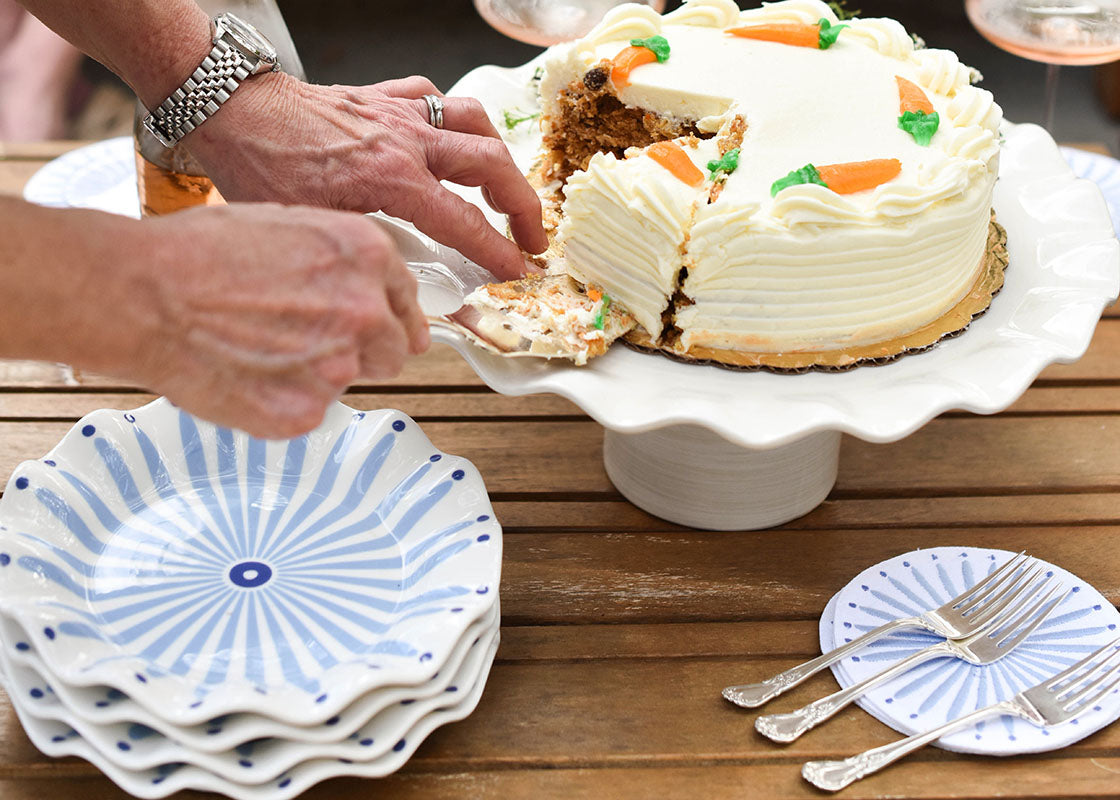 Front View of Hands Lifting a Slice of Cake from Cake Stand with a Stack of Iris Blue Burst Ruffle Salad Plates Set of 4 Placed on the Side