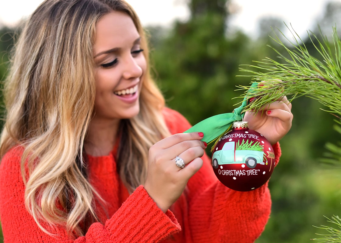 Front View of Woman Tying O Christmas Tree Farm Ornament on Christmas Tree with Green Bow