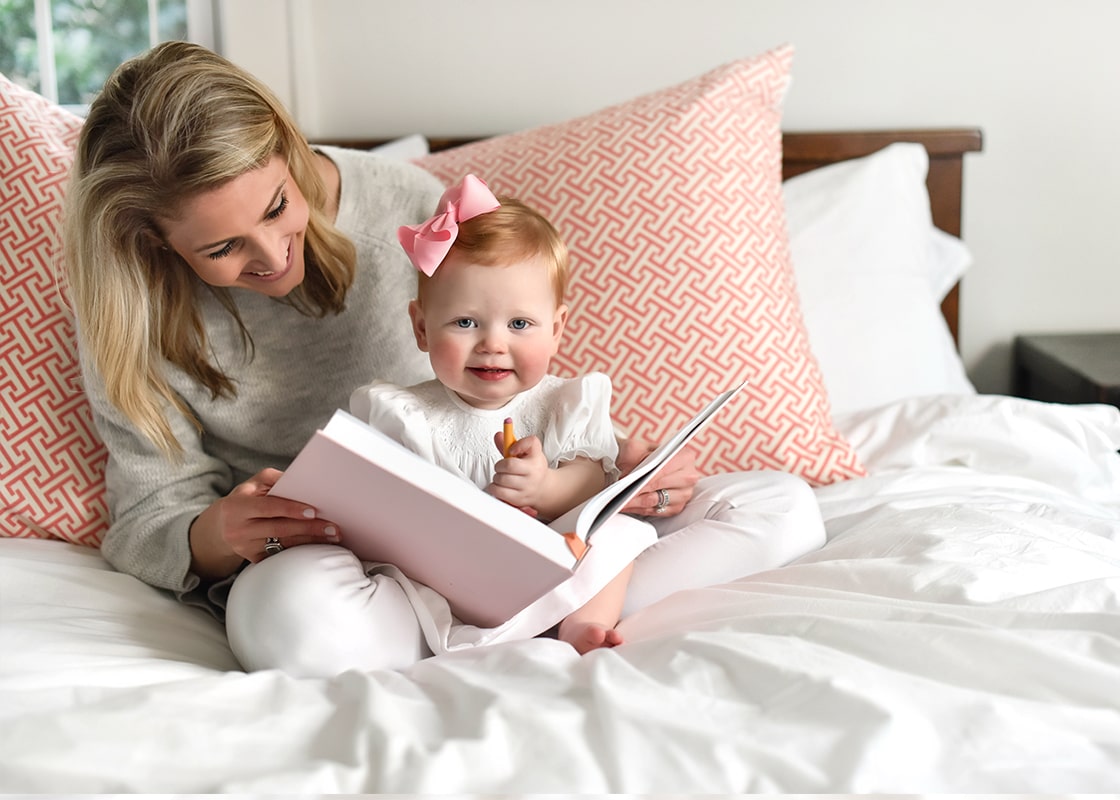 Front View of Sweet Woman Smiling and Sitting on bed with Child on Lap Holding Open Pink Celebrate Me Book