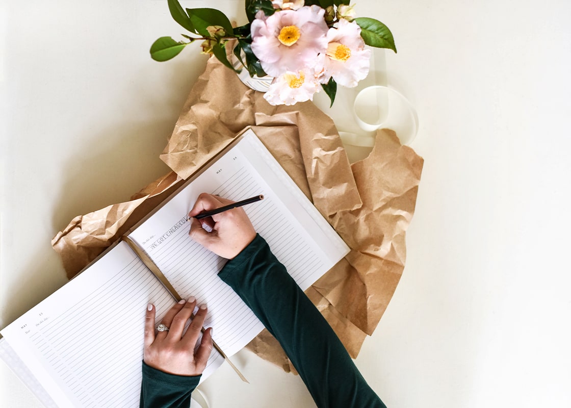 Overhead View of Person Writing We're Engaged Story in Open Celebrate Us Book Surrounded by Gift Wrapping and Flowers