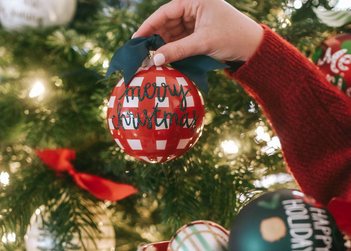 Front View of Person Holding Merry Christmas Ornament with Red Check Pattern in front of Green Christmas Tree