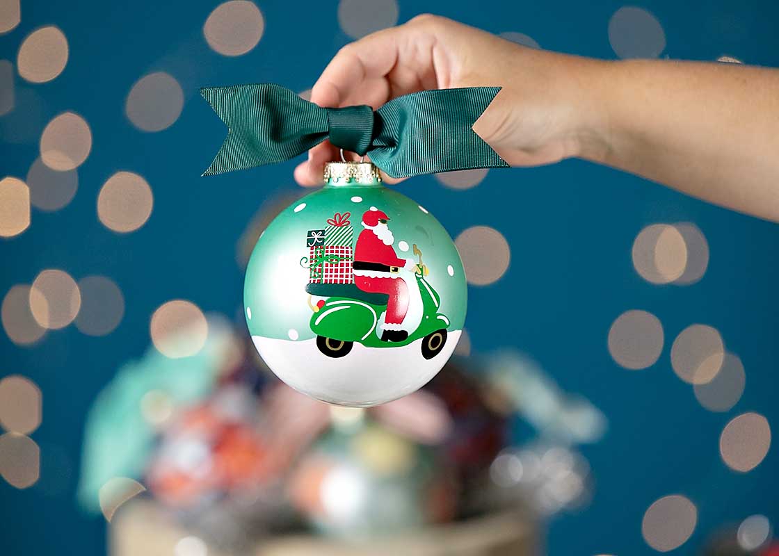 Front View of Hand Holding Up Christmas Ornament Here Comes Santa Claus Against Blue Starry Backdrop