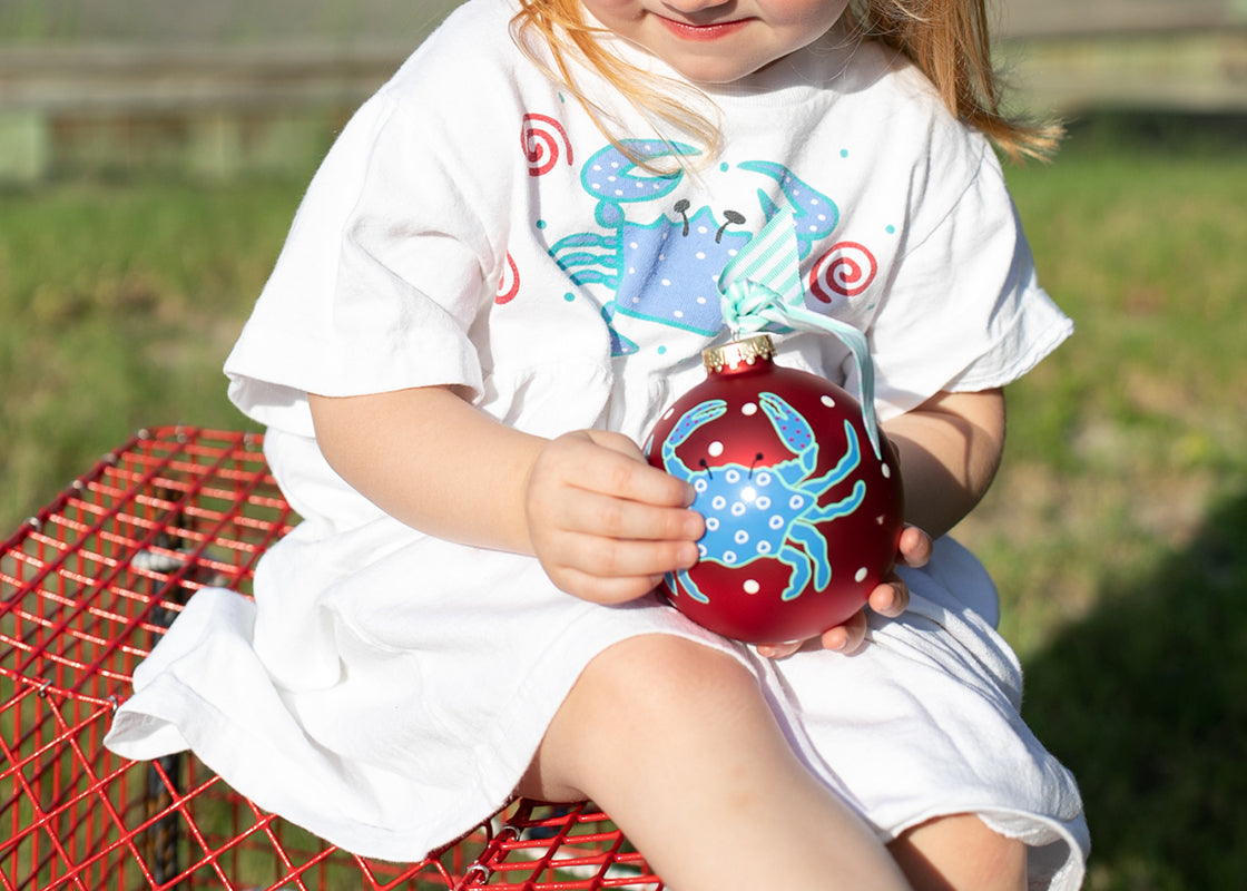 Front View of Child Sitting and Holding Festive Decor Red Ornament Blue Crab Design with Both Hands