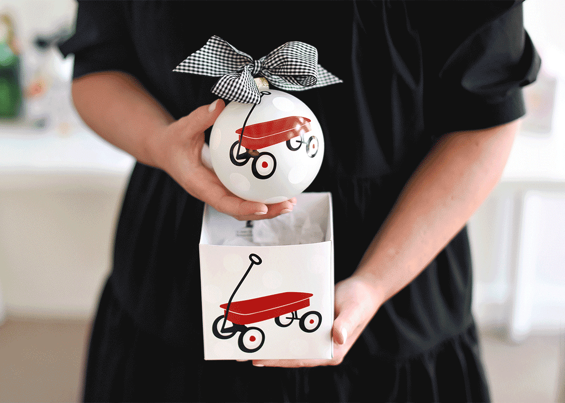 Front and Back View of Woman Holding Red Wagon Ornament in one Hand and Matching Box in the Other Hand Turning Ornament to Show Front and Back