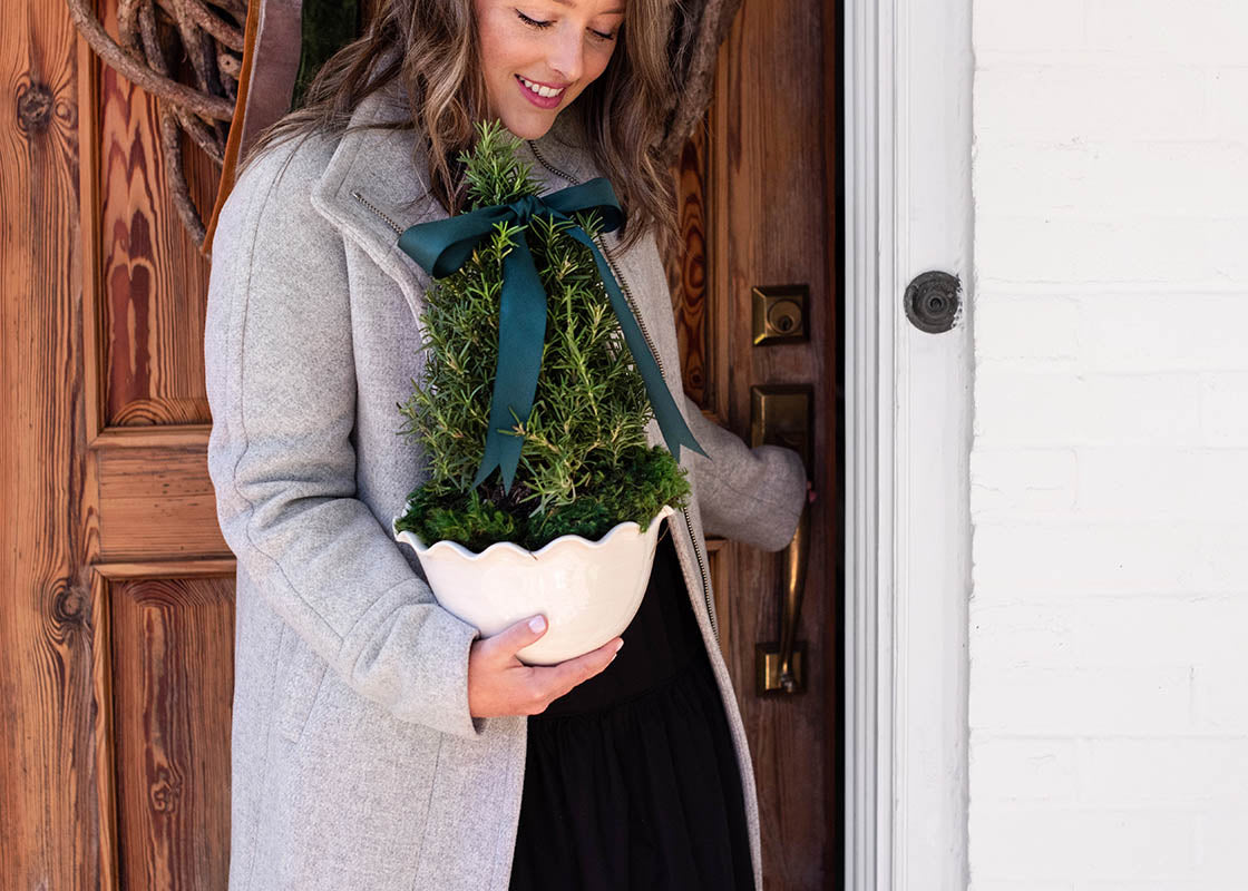 Front View of Woman Standing in Front of Door Holding Small Tree in Signature White Ruffle Bowl Used as Planter